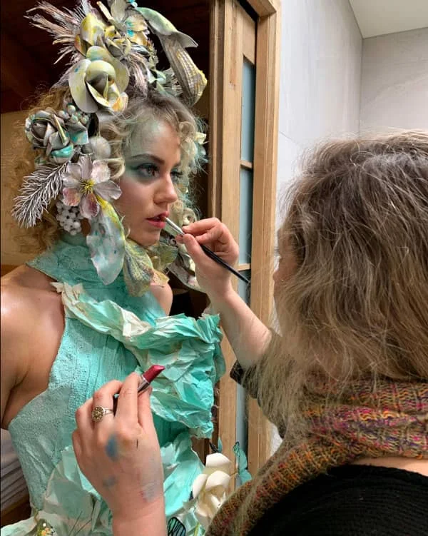 makeup artist applying lipcolor to face of young female model dressed in aqua paper outfit and paper flower headpiece