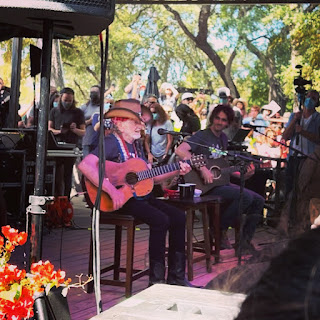 Willie Nelson, hair braided, straw cowboy hat on, sits on a stool playing guitar. A band surrounds him.