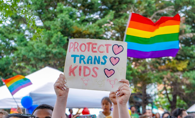 Sullo sfondo bandiere arcobaleno. In primo piano un cartello, con la scritta: "protect our trans kids"