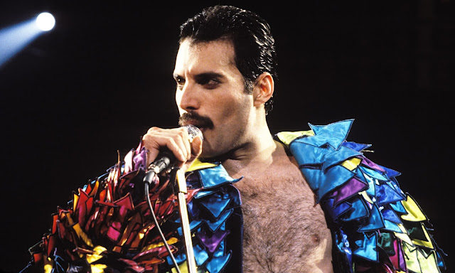 Na imagem: Freddie Mercury segurando o microfone e olhando para o horizonte, vestido de uma camisa espalhafatosa toda colorida e aberta deixando o peito exposto. 