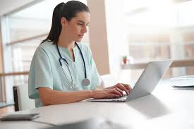 A female trainee nurse in light green nurses uniform, with a doctor's instrument around her neck, studying through continuing education, using a grey laptop on a table..