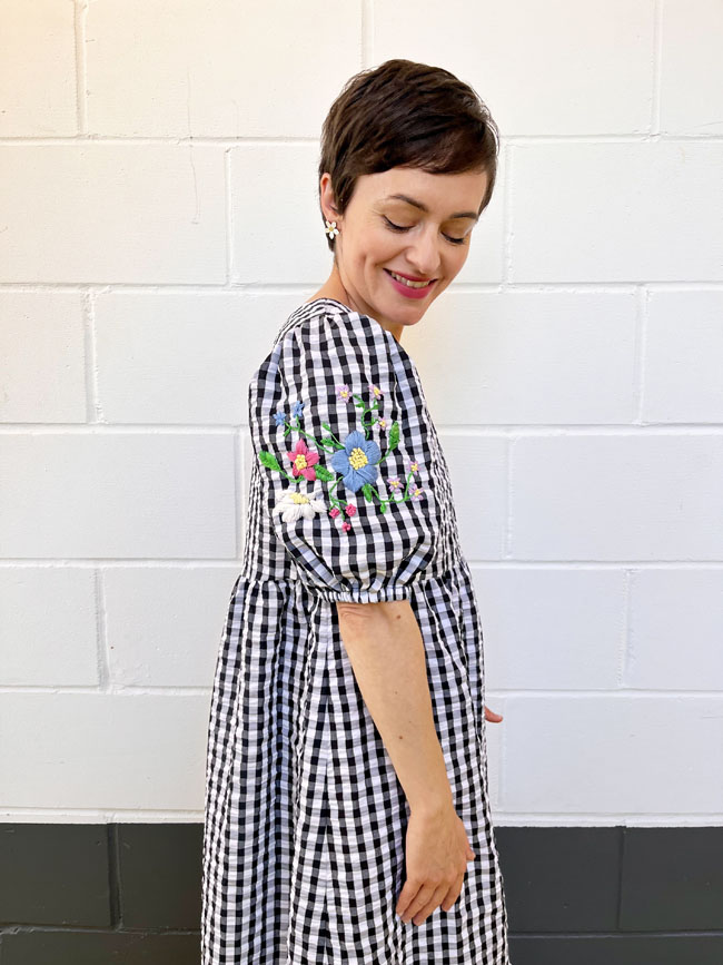 Tilly wears a black and white gingham midi dress with hand-embroidered flowers on the puff sleeves