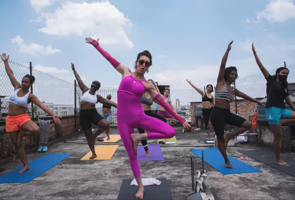 Uma mulher ensina outras a fazer exercícios físicos numa laje. Elas vestem roupas apropriadas para a ação e estão com os braços abertos, o corpo levemente inclinado para o lado esquerdo e uma perna se apoia à outra.