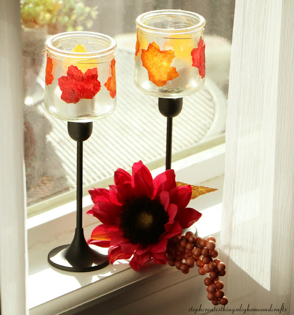Glass jars decorated with faux fall leaves and gold wire attached to taper candle holder displayed on windowsill near a purple flower