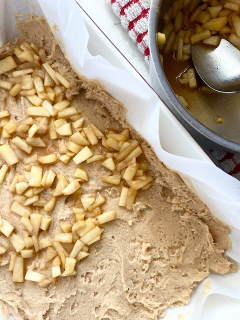 Spiced apples laying over blondie batter with stainless steel bowl of apples and spoon.