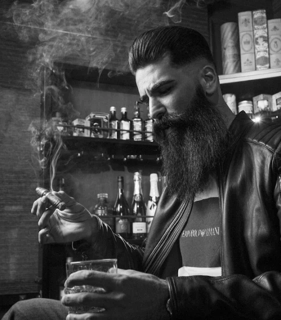 Black and white of long bearded hipster smoking stogie with scotch shot glass in hand sideview