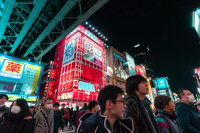 Locals, tourists and otaku walking and shopping around Akihabara district near SEGA red building full of anime billboards at night.