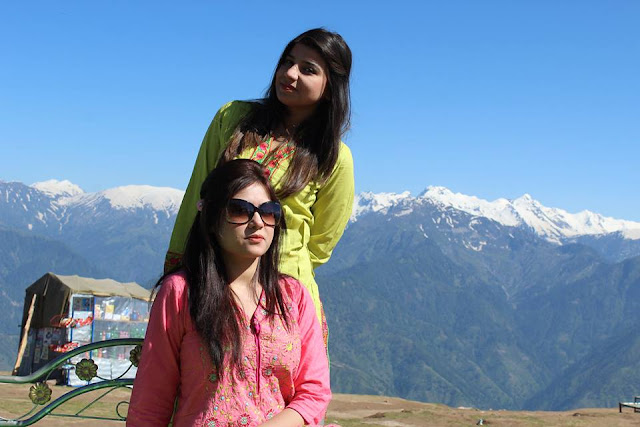 A Girl In Green Standing In Front Of The Majestic Mountains Covered In Snow, With another Girl In Pink