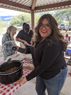 A woman with long, dark, wavy hair wearing glasses and a long-sleeved black shirt. She is holding the lid of a crockpot. The black crockpot is sitting on a table with a red and white checked tablecloth.