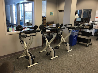 Row of three stationary bikes with laptop workstations arranged against a wall in an office-building setting