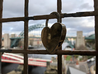 A photo of a padlock on a metal grill on the High Level Bridge.  The padlock is gold in colour and shaped like a love heart.  In the background is the Tyne Bridge.  Photograph by Kevin Nosferatu for The Skulferatu Project.