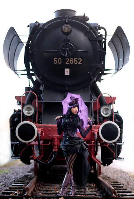 woman dressed in steampunk gothic victorian clothing in purple and black steamgoth style standing in front of a steam train holding an umbrella on railroad tracks