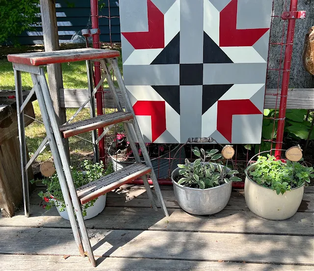 Photo of potted herbs, barn quilt & small rustic stepladder on the deck.