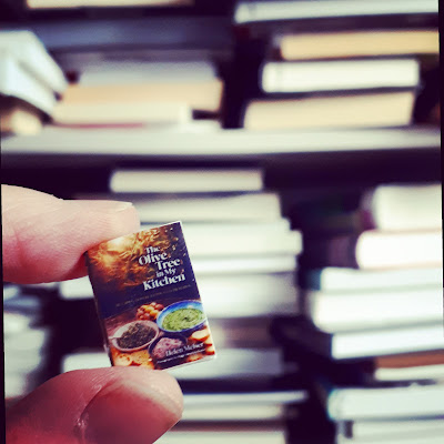 Fingers holding a one-twelfth scale cookbook in front of a bookcase stacked with full-sized books.