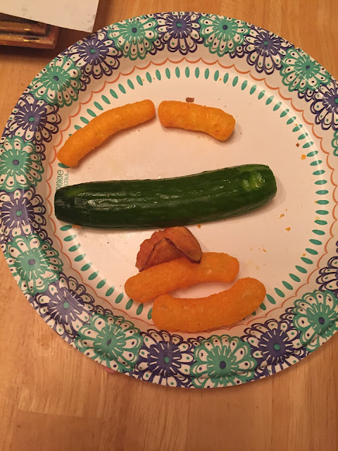 A photo of a plate with a face made from Cheez Doodles, potato chip, and cucumber.