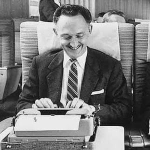 black and white photo of a mustached guy in a suit smiling and typing on an old-style typewriter