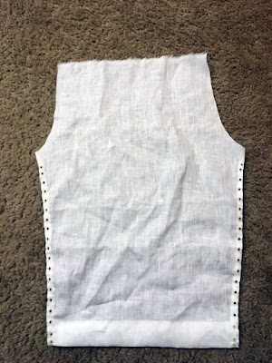 A roughly trapezoidal piece of white linen, with a deep bottom hem, narrow side hems with columns of small eyelets in them, a frayed top edge, and curved cutouts at the upper corners, laid flat on a beige carpet.