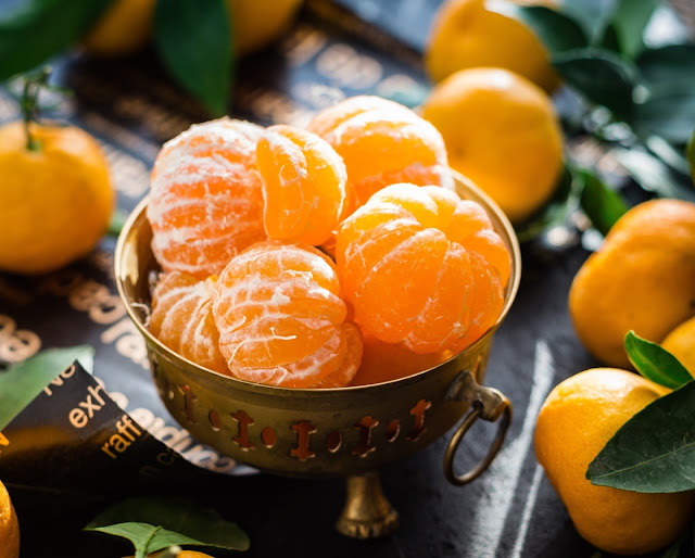 Many peeled oranges in a brass gold bowl and numerous unpeeled ones outside of it with a lot of green leaves.