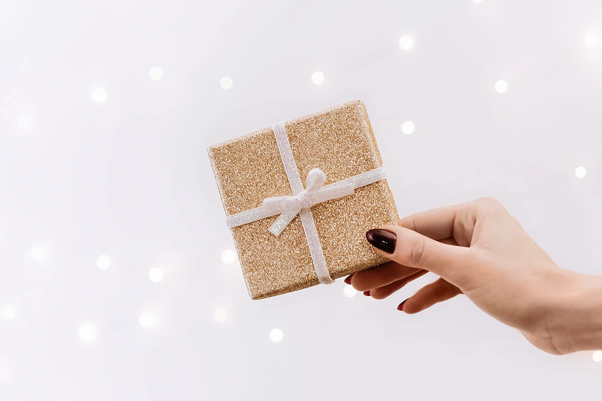 female hand with bright red manicure is holding a small christmas gift box wrapped in sparkling paper