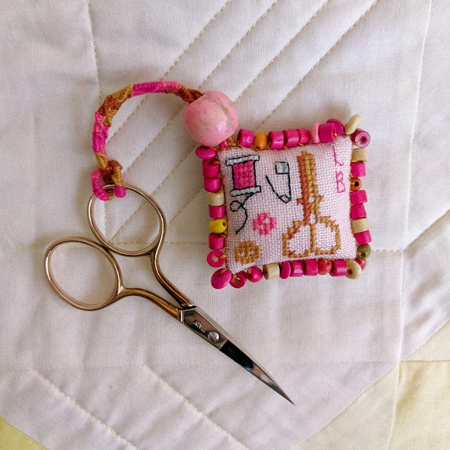 cross stitched scissor fob with wooden beads around the edges attached to pair of scissors, the cross stitch is of scissors a spool buttons and a safety pin along with initials LB