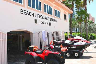 The beach lifeguard station, whose name is displayed in black letters. Underneath, it says: "Tower 0." It is painted a light peach color In front of the building is a red four-wheeler, which has a red medical bag behind the seat. Behind the four-wheeler is a white, red, and black jet ski that is on a trailer.