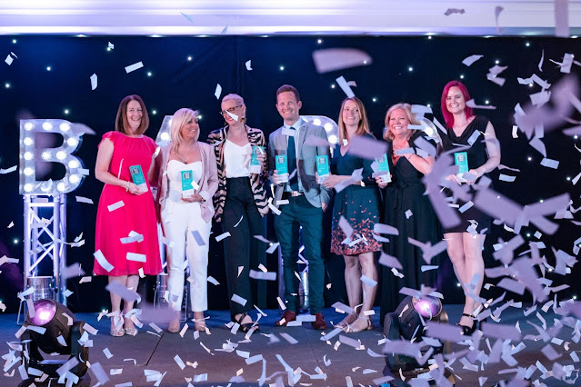 Photo of 7 bloggers on a stage, holding awards, with tickertape floating around everywhere. Steph is far left in a pink dress