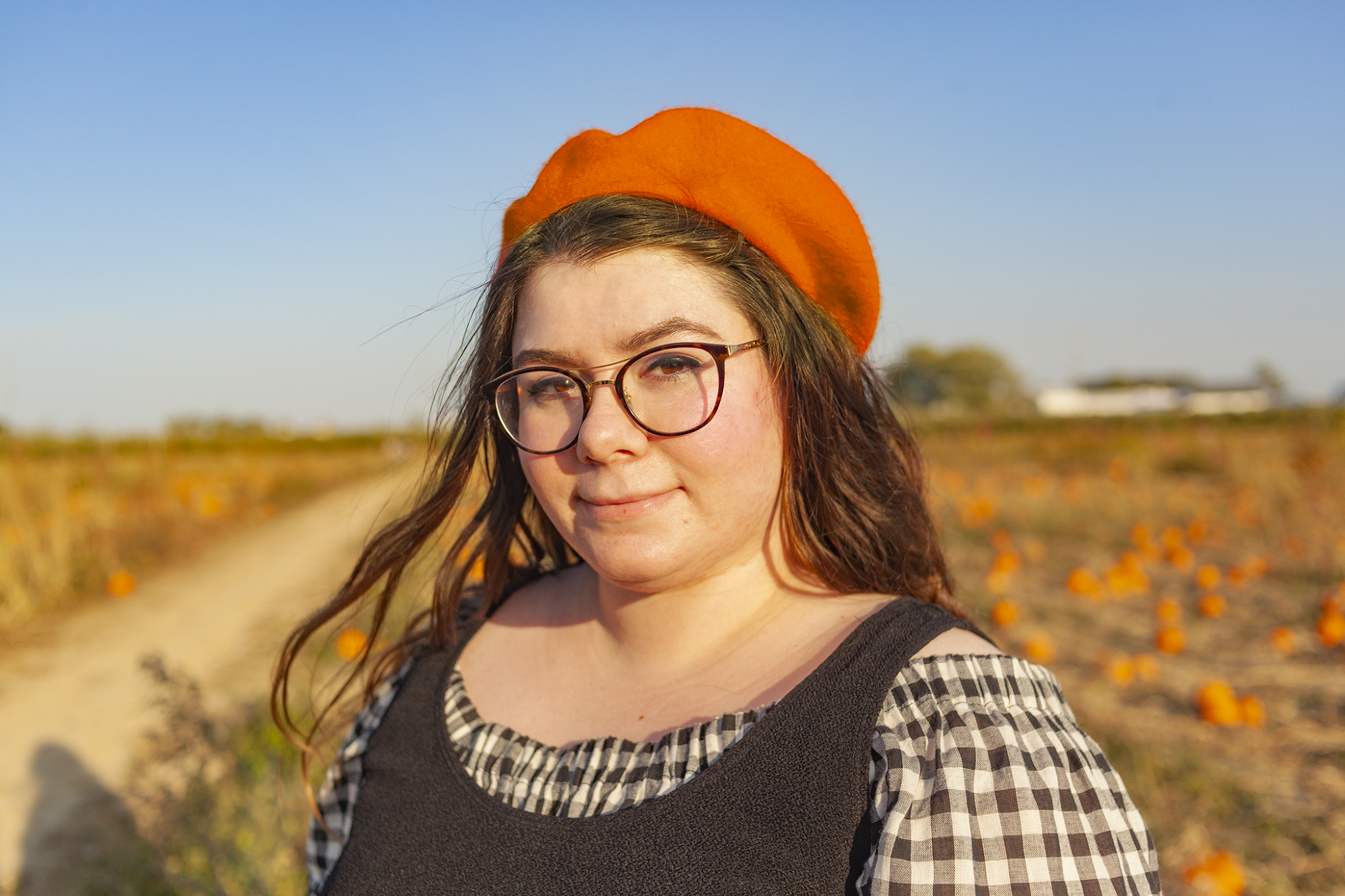 An portrait consisting of a brunette women with a burnt orange beret, wearing a black and white gingham off the shoulder blouse under a sleeveless scoopneck black dress under a black pleated midi skirt. Her eyes are squinting from the sun as she faces the camera on a 3/4 angle.