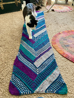 a black and white cat walks along a large, long knit wrap stretched across a floor