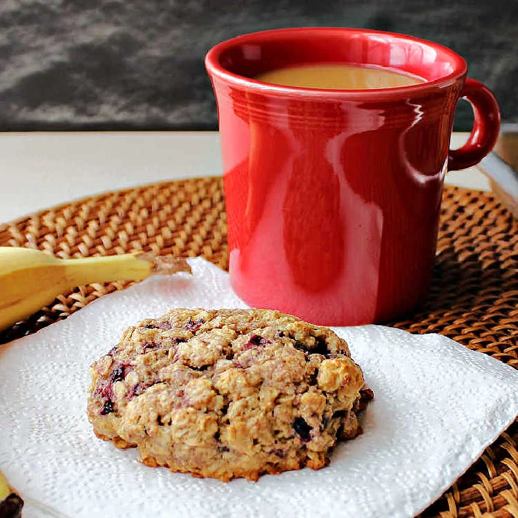 A large black berry oatmeal breakfast cookie on a napkin with a cup of coffee