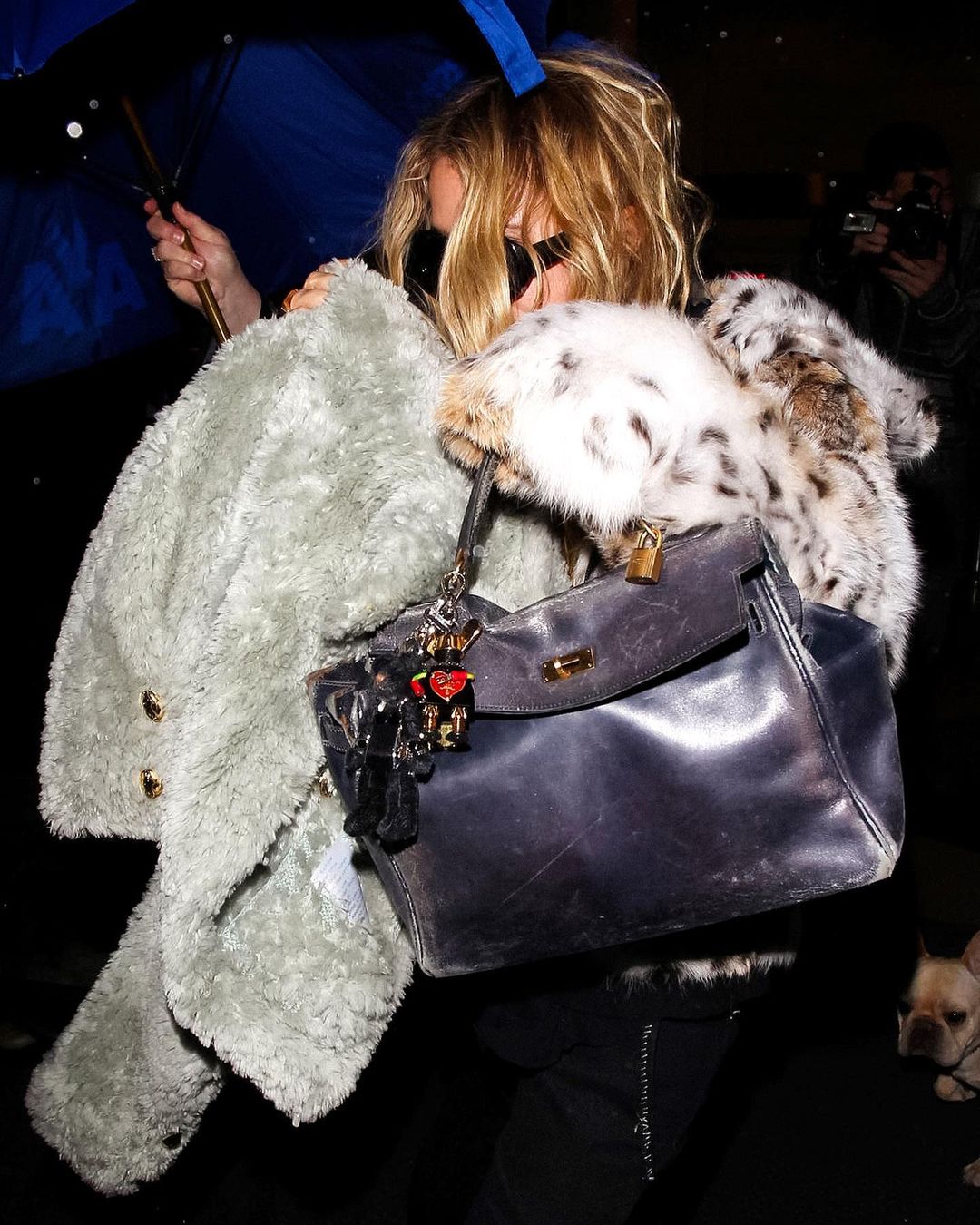 Woman with blonde wavy hair and black oversized round sunglasses wearing a fur mink jacket with a hood and black pants with silver zipper details carrying a grey fuzzy sweater with gold buttons, a royal blue umbrella, and a navy blue oversized Hermes Birkin bag with gold lock and keychain charms