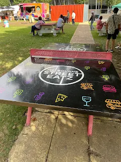 Table tennis table. Top of the table is black with the logo "Melbourne Street Eats" and line drawings of hamburgers, hotdogs, and french fries in bright neon pink, yellow, and blue. The edges, legs, and net of the ping pong table are bright pink.