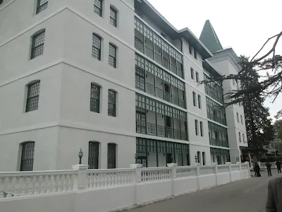 Hotel Cecil colonial-style white building with green window frames and a sloped green roof, surrounded by a white fence and tall trees, located in Shimla hill station.