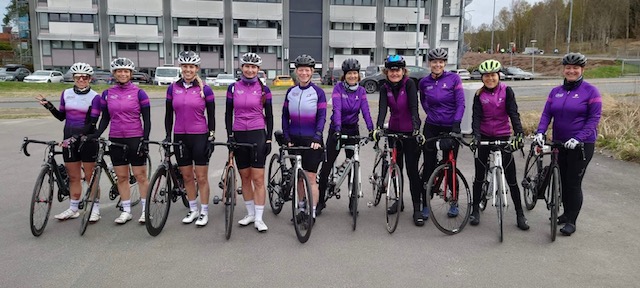 Gruppebilde av 10 damer som står i sykkelutstyr ved sykkelene sine.