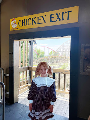 A light-skinned, blond girl in a blue dress with white lace, stands in front of a doorway with a rollercoaster in the background. Above the door is a sign that says "Chicken Exit."