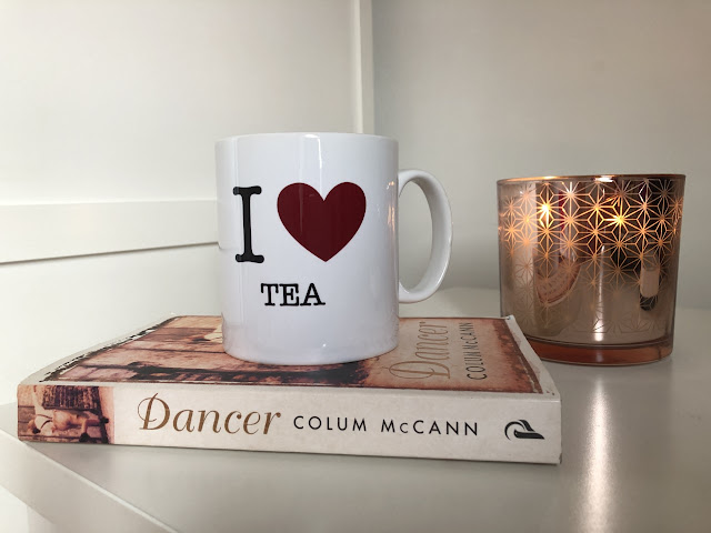 Dancer by Colum Mccann book with I Love Tea mug on top next to rose gold candle