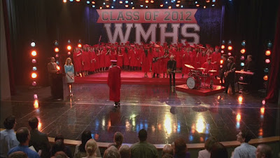 Graduation stage with a sign that says Class of 2012. A bunch of kids on stage are wearing red graduation gowns and caps and there is a band on the stage