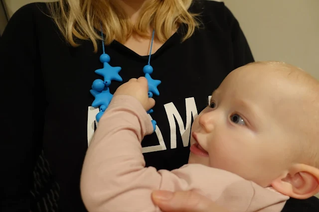 A baby holding on to a blue teething necklace with blue stars and balls, while being held