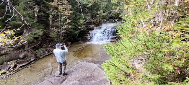 Man standing on a boulder and taking a picture of a waterfall.