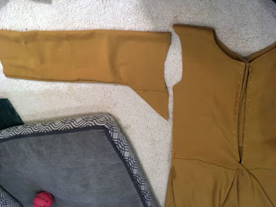 A photo from above of about three-quarters of a muddy-gold dress bodice, which runs out of frame at right and bottom, and has a shallow scooped neckline and armscye. The corresponding sleeve runs horizontally left from the bodice, with a small triangle of fabric pointing down at the armpit. A thick grey dog bed fills the lower left corner of the photo.