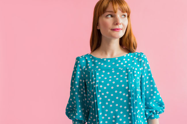 A red haired model wears a teal polka dot Mathilde blouse with puffed sleeves and tucks down the bodice