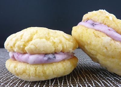 Two lemon cookies sandwiched together with blueberry buttercream, photographed on a grey woven mat