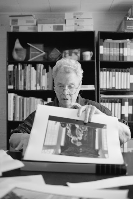 Joan E. Biren, já com idade mais avançada e cabelos grisalhos, sentada em um ambiente que parece ser uma biblioteca e olhando um livro de fotografias. Ela está com o semblante sério e atento, o que passa a impressão de que procura alguma imagem específica.