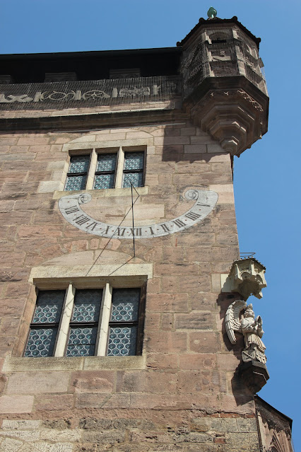 A sundial painted in white on a medieval stone tower that has the stone statue of an angel on its corner..