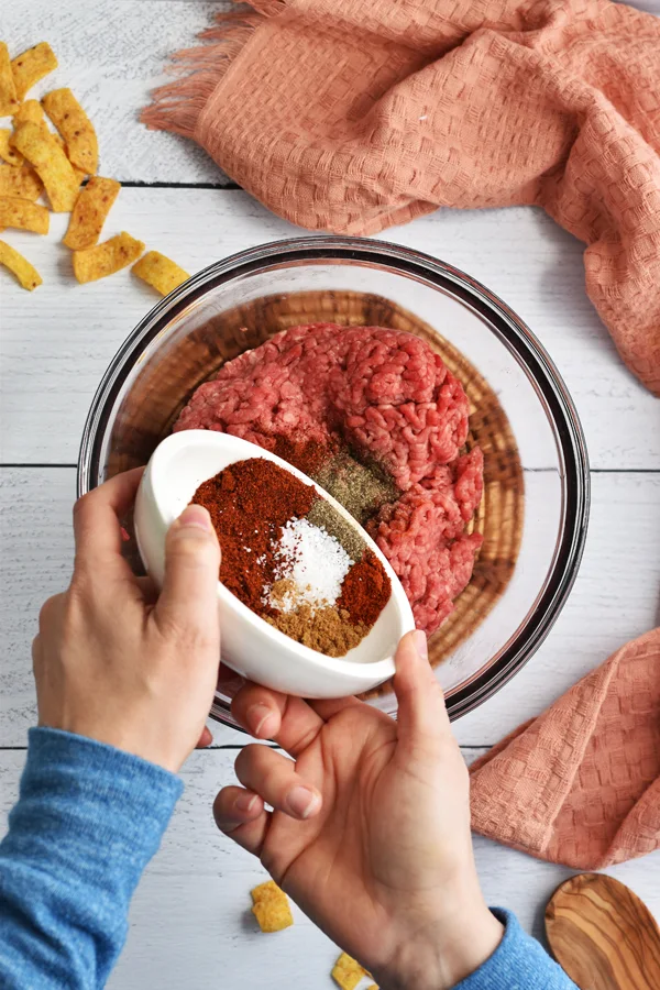 Hands holding a white bowl with seasonings and pouring the seasonings into the mixing bowl with the ground beef