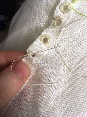 A close-up of a white finger and thumb supporting the narrow double-folded hem of a piece of white linen, with a needle inserted through the small round hole and tucked over the working thread, ready to form the next buttonhole stitch. Thickly worked eyelets follow the hem to the right, and a small scrap of bright green thread is just visible at top right.