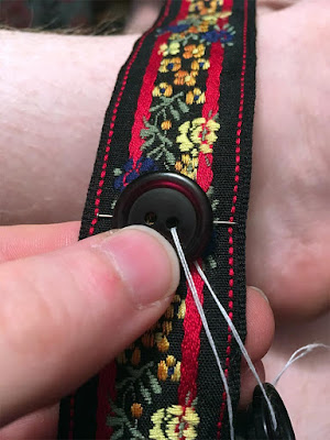 A white hand holding a dark, translucently red button against a floral-patterened ribbon. The ribbon is black, with a pair of vertical red stripes and yellow, orange, and blue flowers overlaid. A pin marks the spot for the button, which has a doubled white thread running through it to other buttons below.