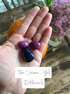 a walnut stained table holding an orange pumpkin and bright pink flowers in a vase all behind the palm of a hand which is holding three beans, one dark-purple nearly black, and two purple-pink ones