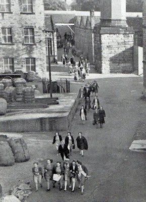 A black and white photograph showing several groups of women walking between mill buildings