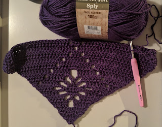 Work in progress crochet triangle shawl in purple acrylic yarn, with the ball of yarn above with label "Marvel soft 8 ply". The crochet hook attach at the top of the triangle of crochet.