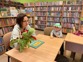 Pani bibliotekarka trzyma pacynkę t-rexa i zadaje dzieciom pytania. Tło: regały z książkami.